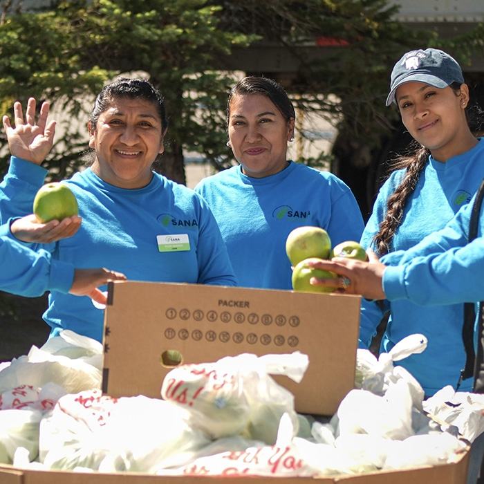 Volunteers gathered around a box of bagged produce, smiling and waving.