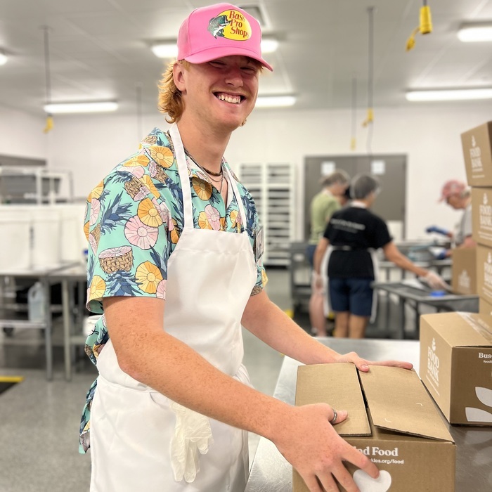 Volunteer in kitchen smiling with hands on a food box.