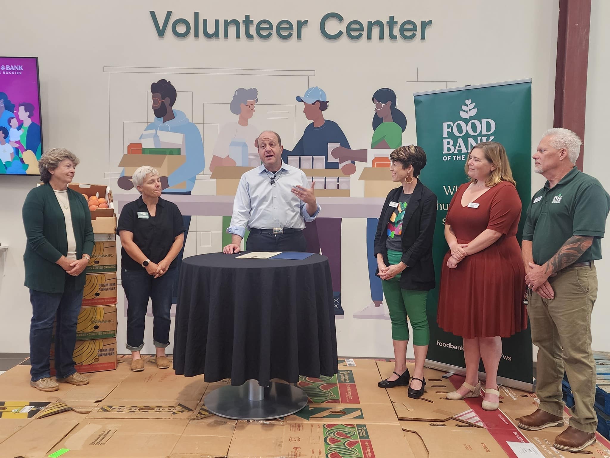 Governor Polis inside the Western Slope distribution center, with a sign that says volunteer center on the back wall. Staff on both sides of him.