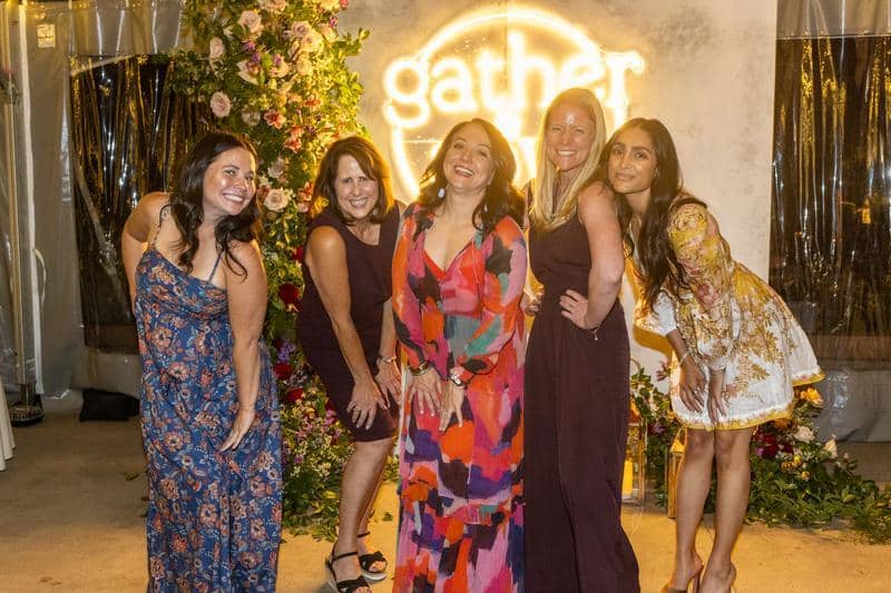 Group of women posing in front of a lit up neon Gather & Sow sign.