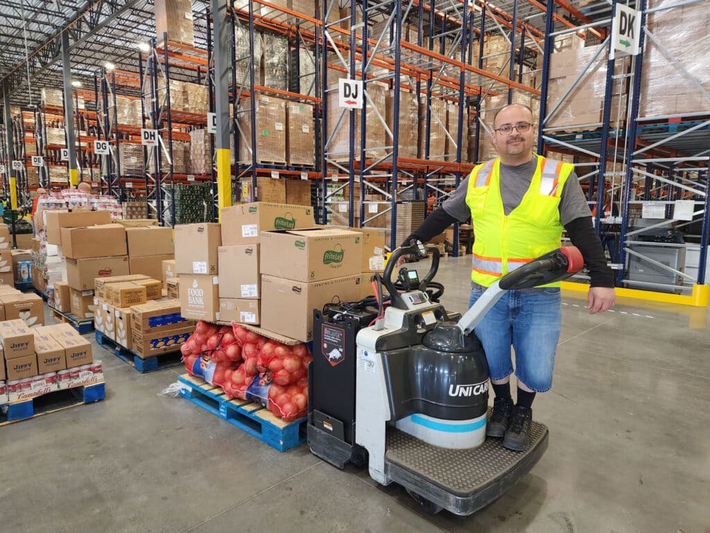 Warehouse worker pulling food boxes and produce on a motorized cart inside a food bank warehouse.