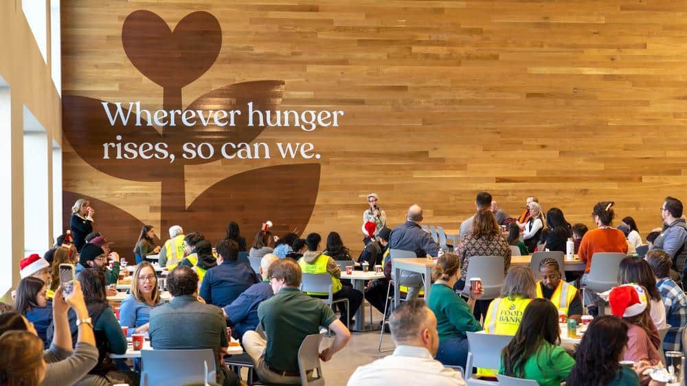 Staff meeting in lobby of distribution center. Wooden panel wall in the back, with text that says: Wherever hunger rises, so can we.