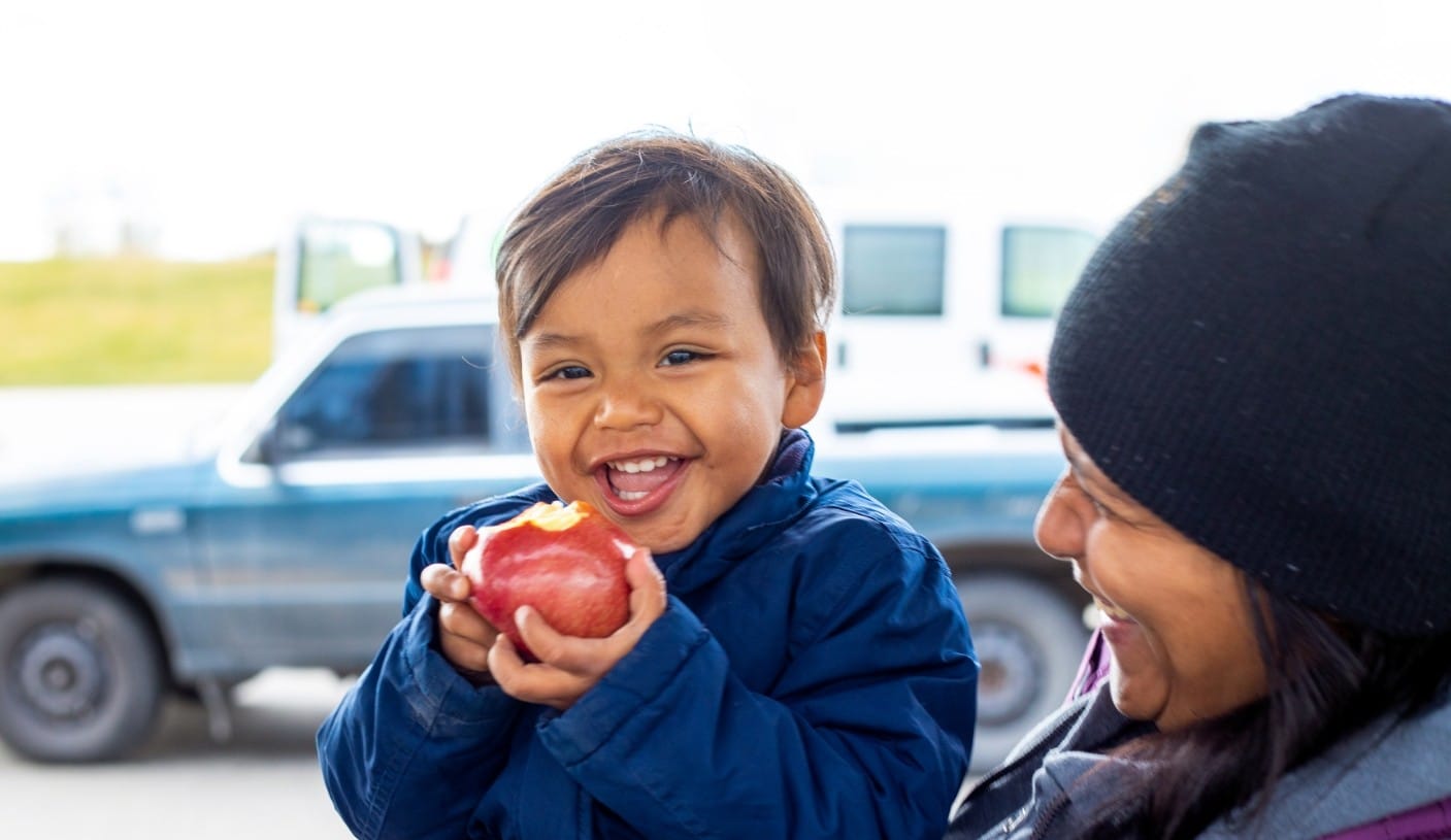 Toddler being held by his mother, holding an apple with one bite already in the apple.